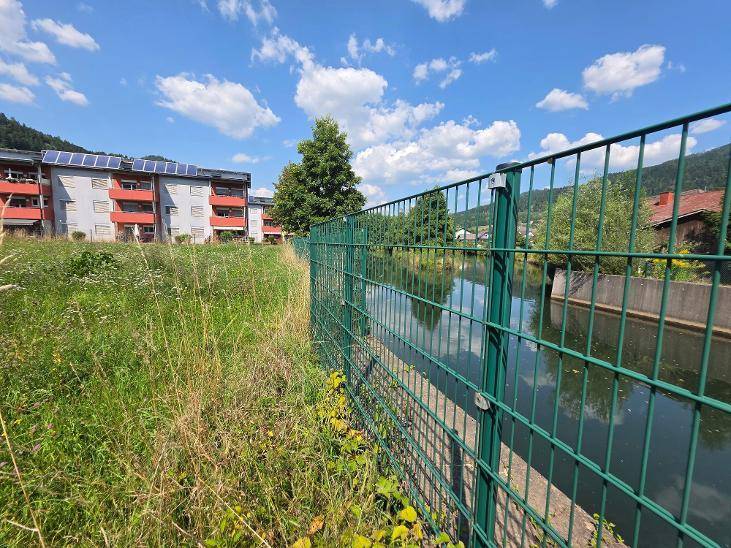 Grüner Metallzaun entlang eines Wasserlaufs mit Wohngebäude und Bergen im Hintergrund.