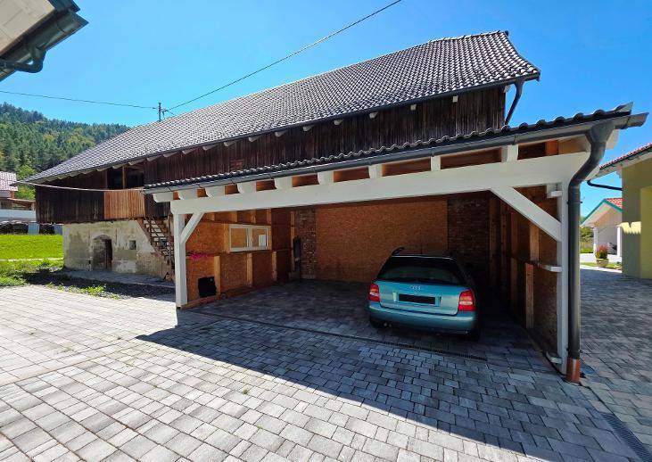 Überdachter Carport neben einem rustikalen Scheunengebäude mit Holzwänden und Ziegeldach.