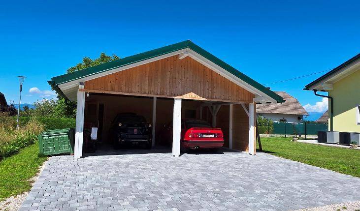 Offener Holzcarport mit grünem Dach bietet Platz für zwei Fahrzeuge auf gepflastertem Untergrund.