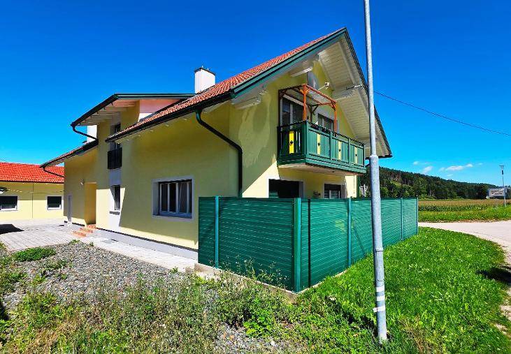 Gelbes Haus mit kleinem Balkon, grüner Sichtschutzwand und Blick auf Felder und Wald.