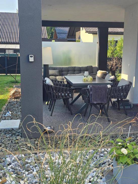 Überdachte Terrasse mit großem Esstisch und bequemen Stühlen, umgeben von Kies und Pflanzen.