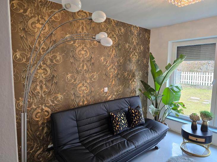 Gemütliches Wohnzimmer mit auffälliger Tapete, schwarzem Sofa und einer großen Zimmerpflanze.