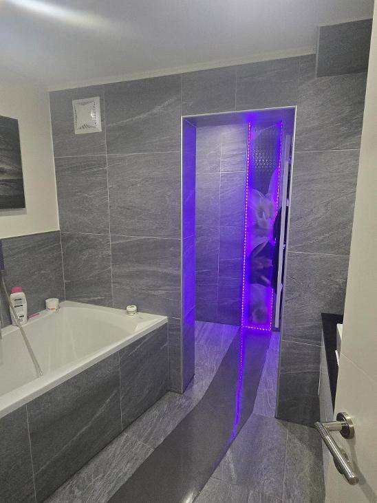 Modernes Badezimmer mit grauen Fliesen, Badewanne und einem beleuchteten Durchgang.