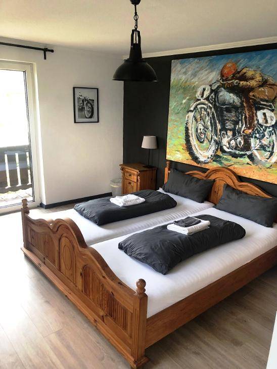 Schlafzimmer mit zwei Einzelbetten, Holzbettrahmen, einem großen Motorradgemälde und Zugang zu einem Balkon.