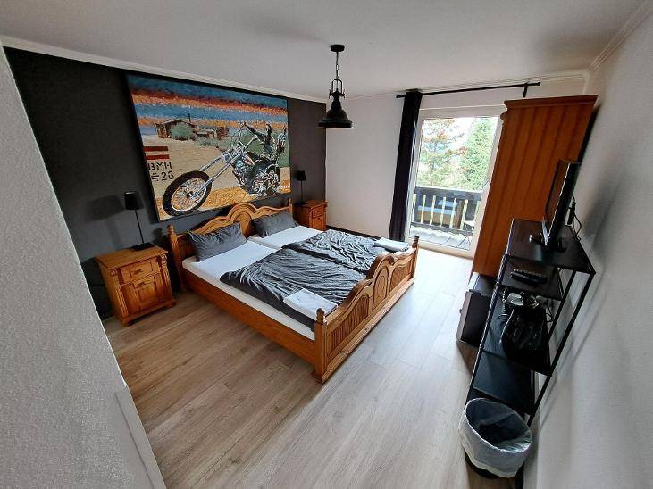 Schlafzimmer mit Doppelbett, Holzbettrahmen, Motorrad-Kunstwerk und Balkonzugang, auf einem Laminatboden.