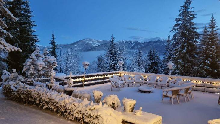 Schneebedeckte Terrasse in der Dämmerung mit Gartenmöbeln, Lichterketten und einem malerischen Blick auf die Berge.