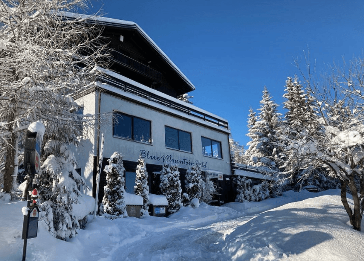 Winteransicht des Gebäudes, komplett mit Schnee bedeckt, umgeben von verschneiten Bäumen unter blauem Himmel.