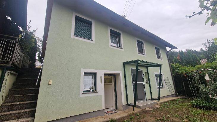 Vorderansicht des Hauses mit grüner Fassade, Rollläden und einem überdachten Eingangsbereich.