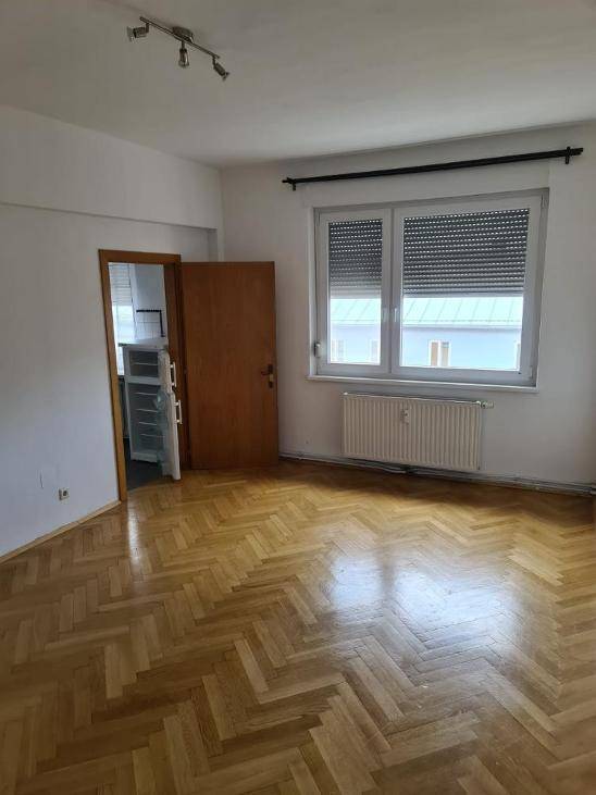 Geräumiger Raum mit Fischgrätparkett, großem Fenster und Zugang zu einer kleinen Küchenzeile.
