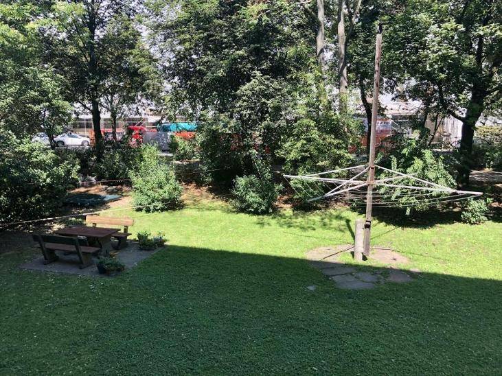 Gemeinschaftsgarten mit Rasenfläche, Picknicktisch und Wäschespinne unter Bäumen.