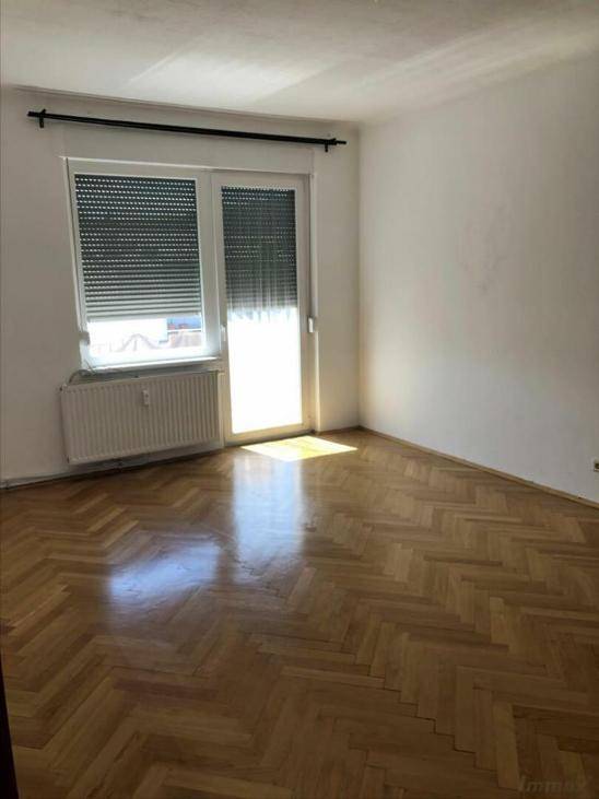 Lichtdurchfluteter Raum mit Fischgrätparkett, Fenster mit Rollladen und Heizkörper.