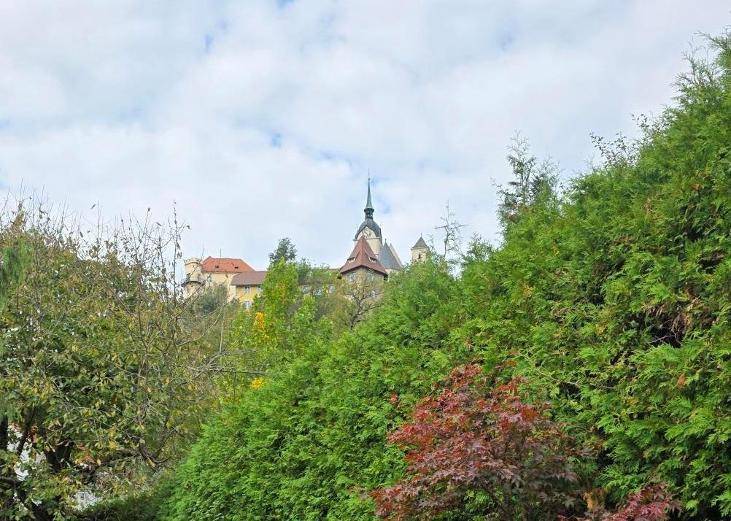 Historisches Gebäude mit Turm, umgeben von dichtem Grün unter bewölktem Himmel.