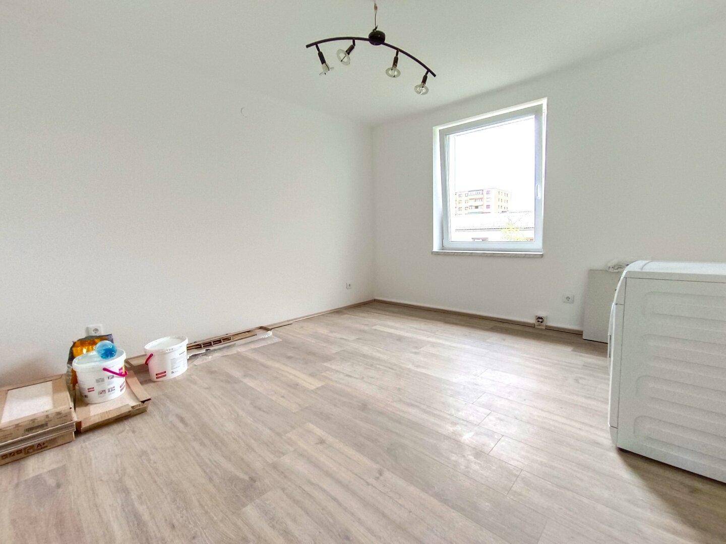 Geräumiger Raum mit hellem Laminatboden, Fenster und Platz für Haushaltsgeräte.