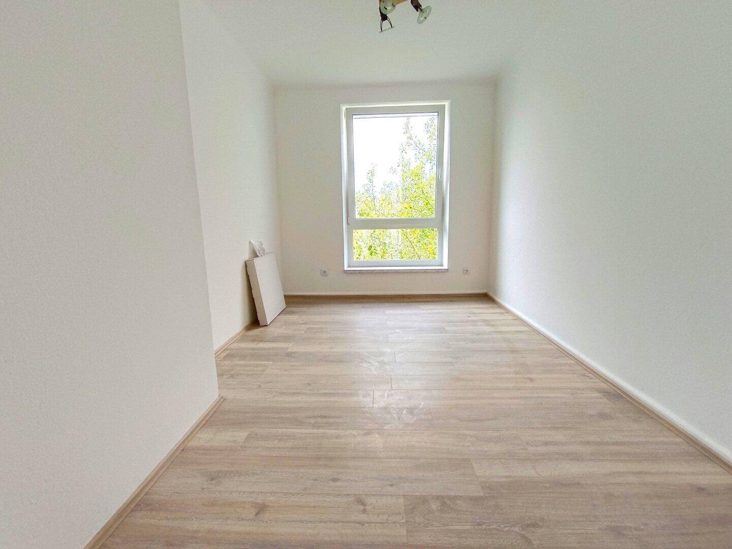 Ruhiges Zimmer mit hellem Laminatboden, weißen Wänden und Fenster mit Blick ins Grüne.
