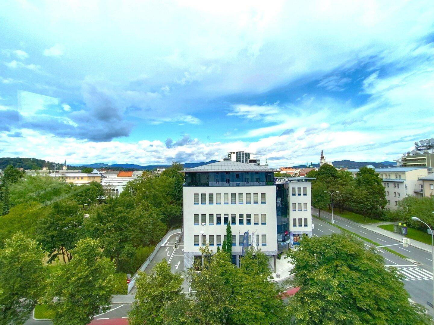 Panoramablick über eine Stadtstraße mit modernen Gebäuden und viel Grün unter bewölktem Himmel.