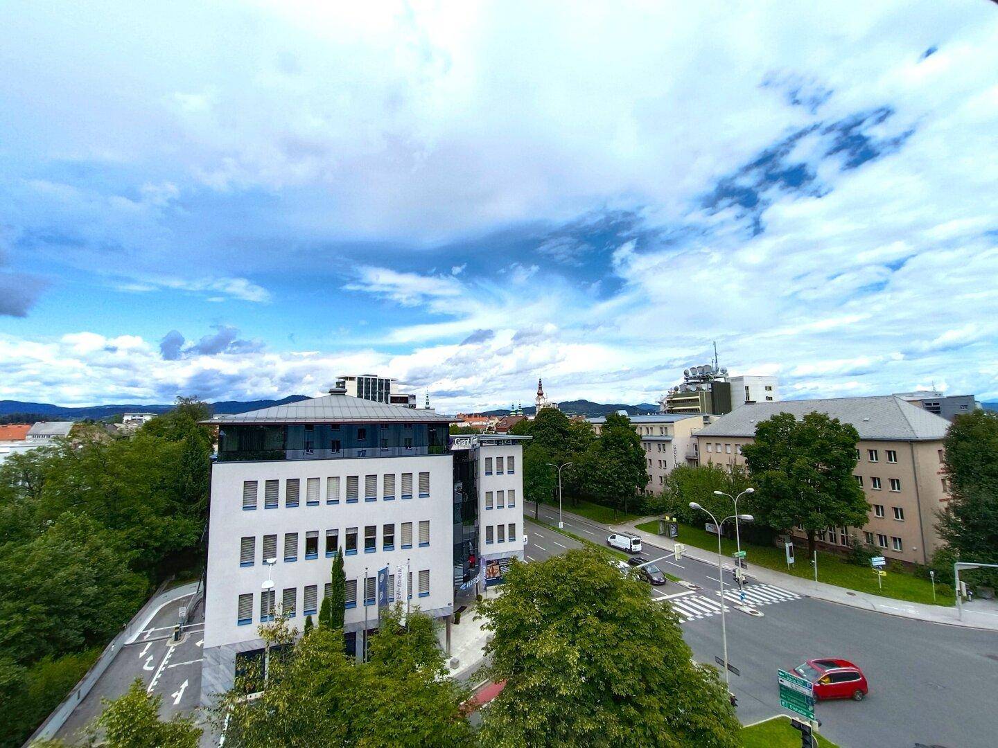Weitläufiger Blick über eine städtische Umgebung mit Gebäuden, Bäumen und einer belebten Straße.