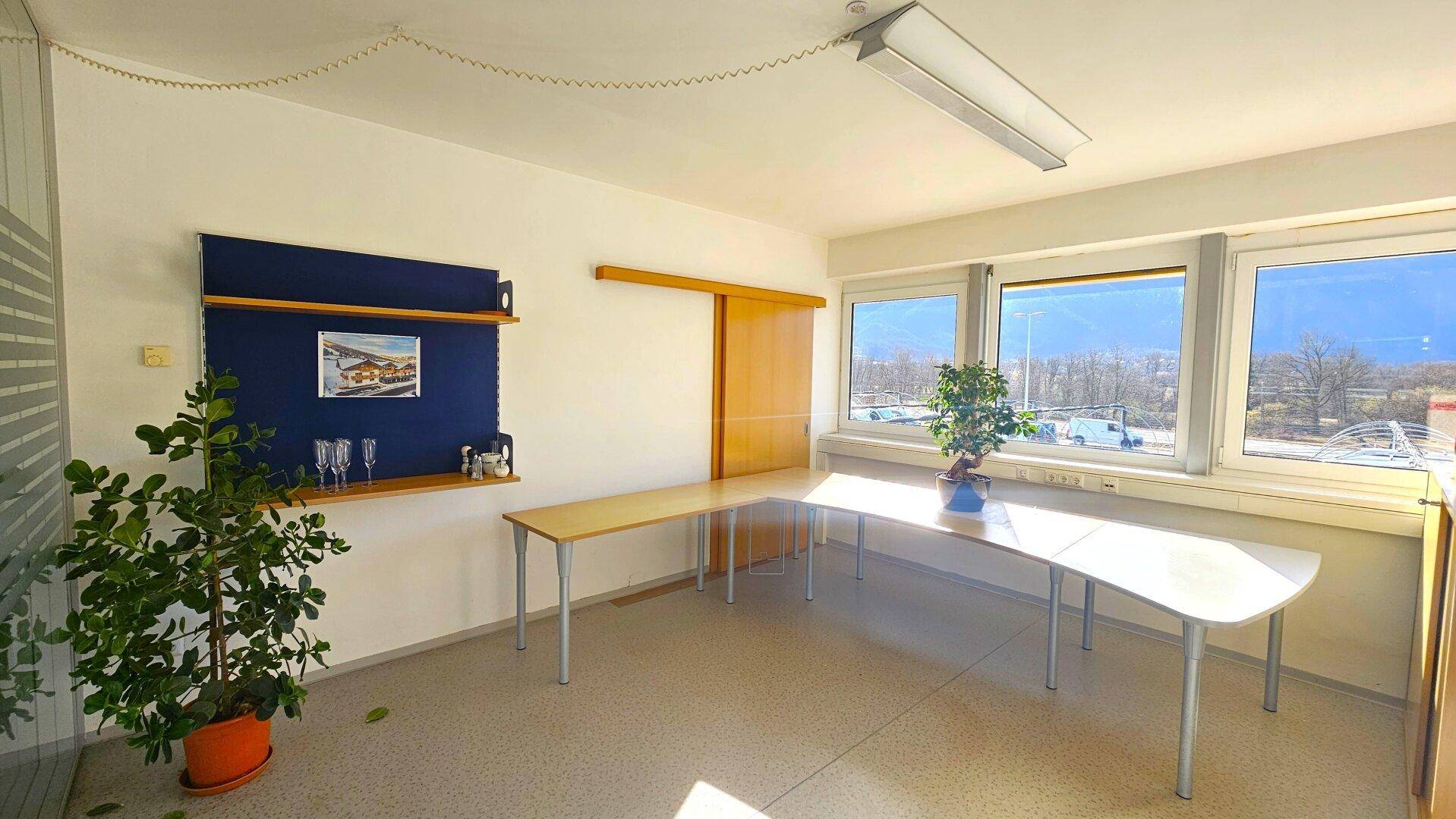Heller Büroraum mit mehreren Fenstern, einem langen Tisch und einer blauen Pinnwand, gut beleuchtet.