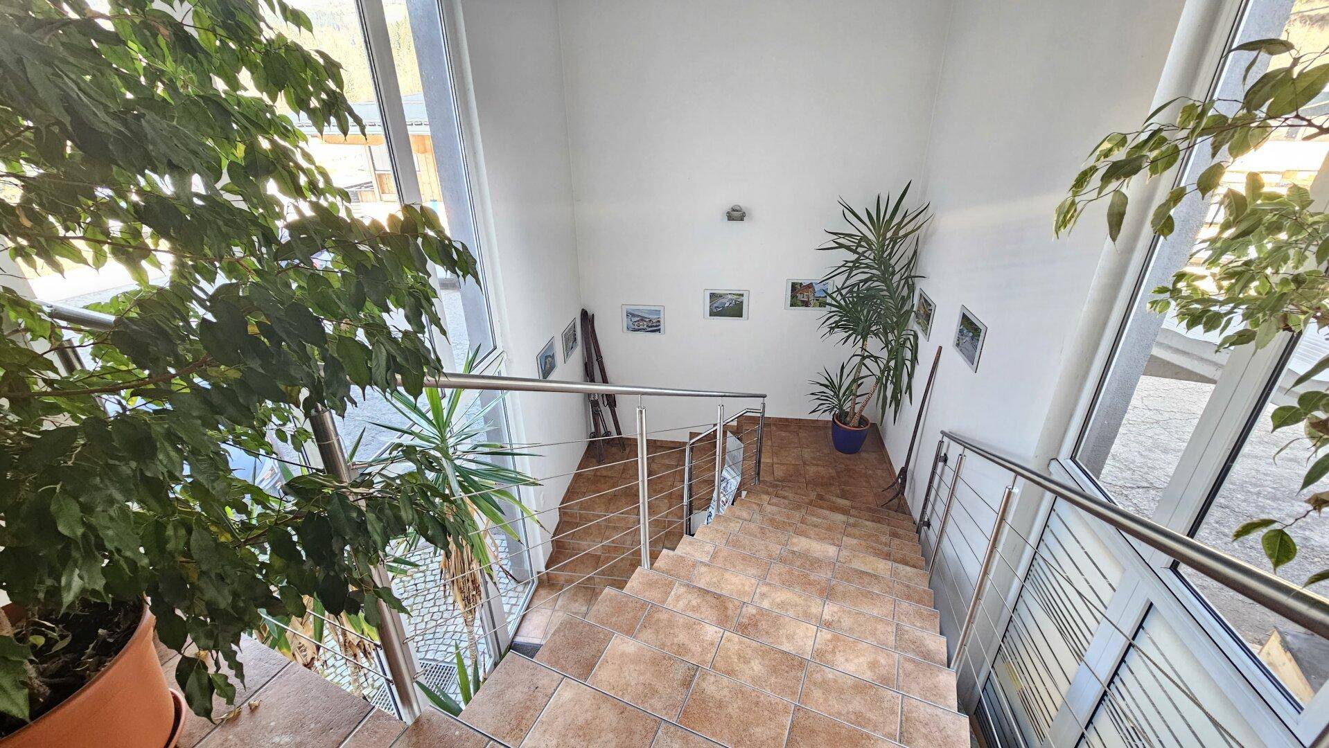 Helles Treppenhaus mit Fliesenstufen, Edelstahlgeländer und großen Fenstern, die viel Licht hereinlassen.