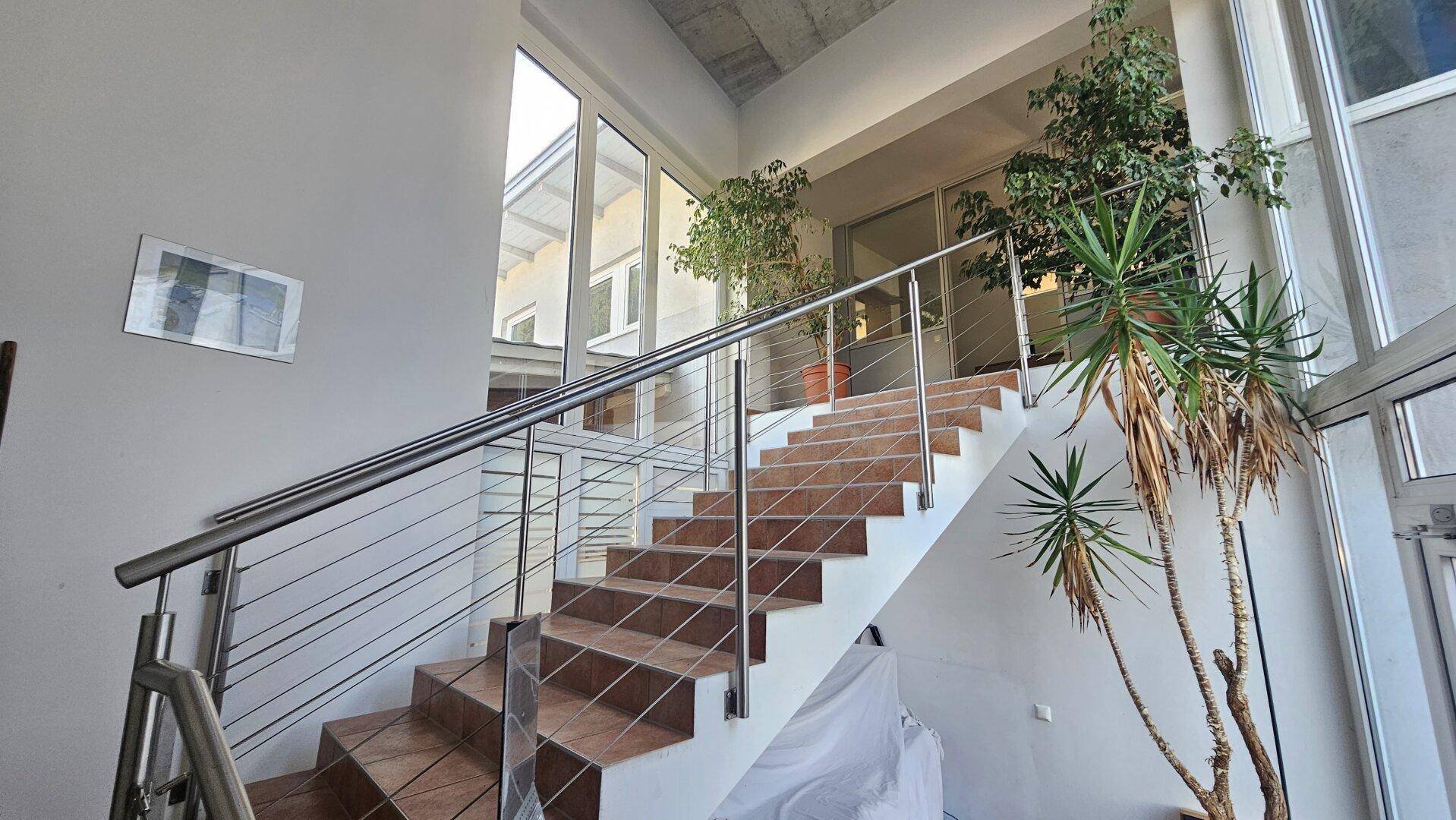 Modernes Treppenhaus mit breiten Fliesenstufen und elegantem Edelstahlgeländer, umgeben von großen Glasflächen.
