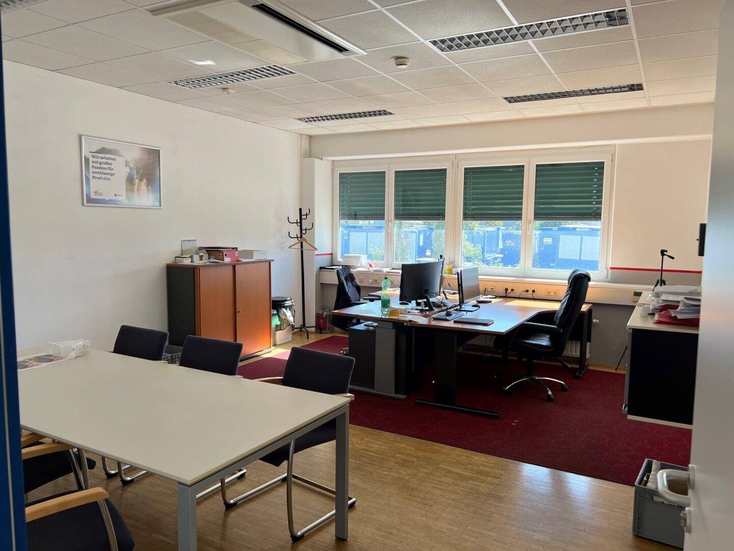 Möbliertes Büro mit Konferenztisch, Schreibtisch und Teppichboden, gut beleuchtet.