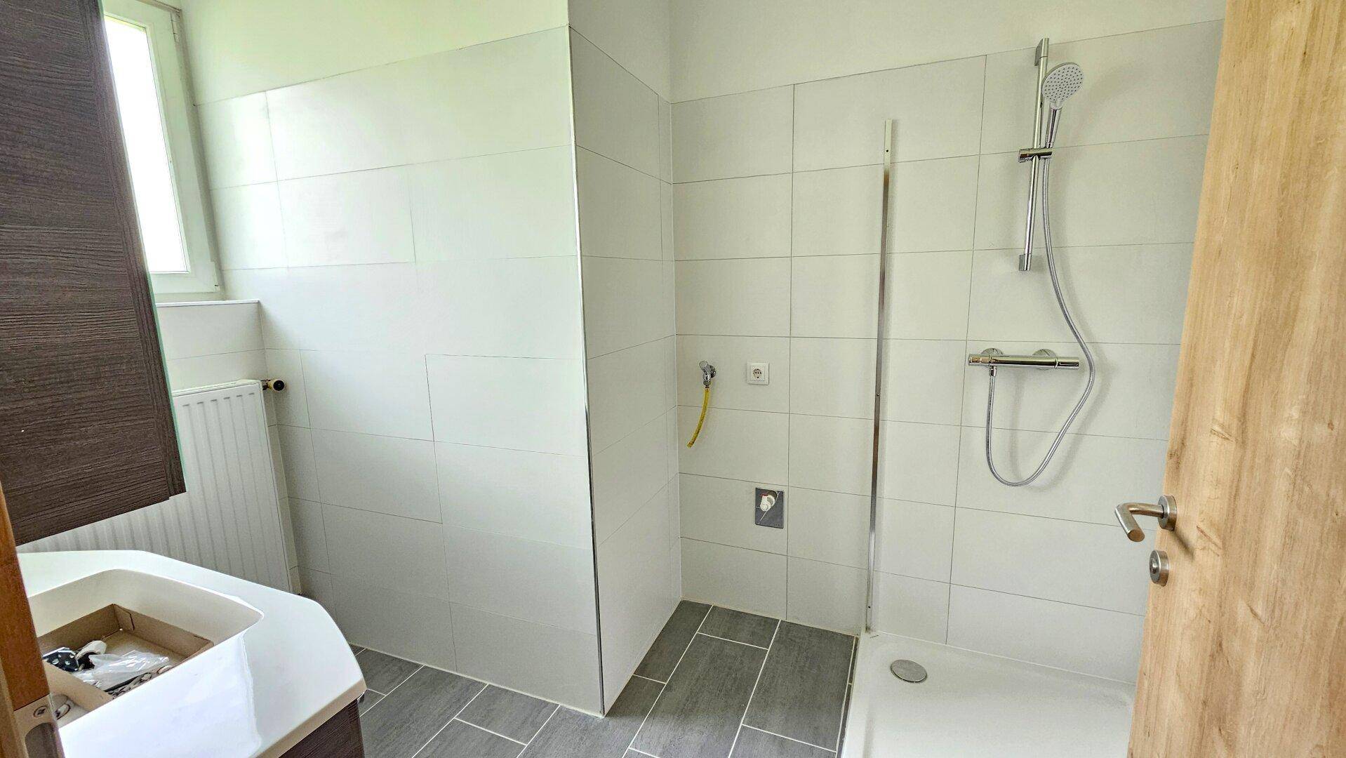 Modernes Badezimmer mit weißen Wandfliesen, grauen Bodenfliesen und einer Duschkabine mit Fenster.