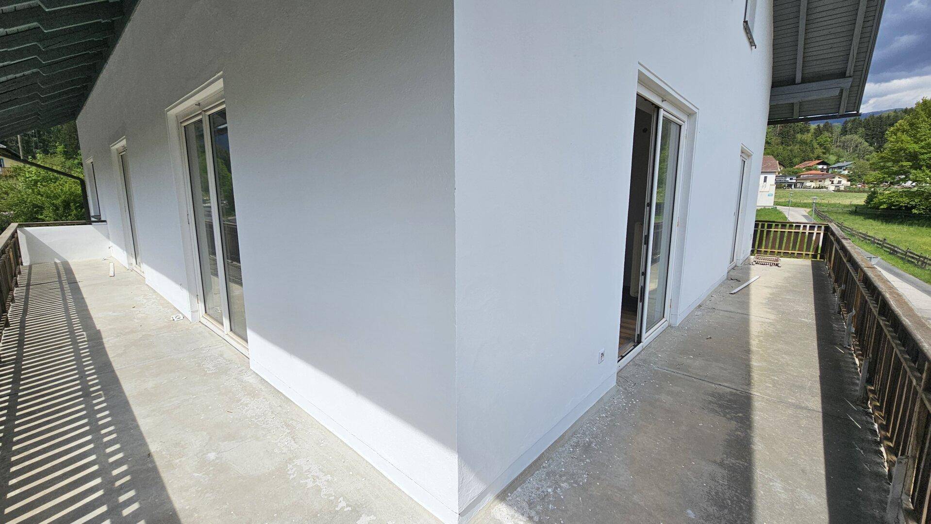 Langer Balkon mit Betonboden und Holzgeländer, der Zugang zu mehreren Räumen des Hauses bietet.