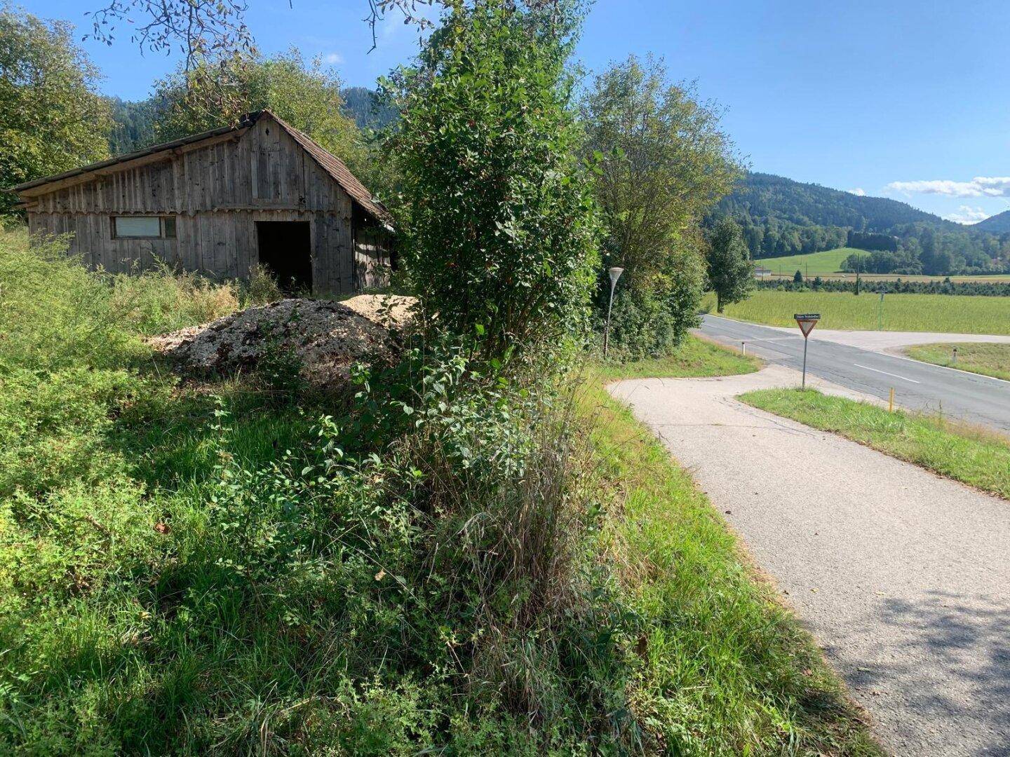 Altes Holzgebäude neben einer Landstraße, umgeben von grüner Landschaft und fernen Hügeln.