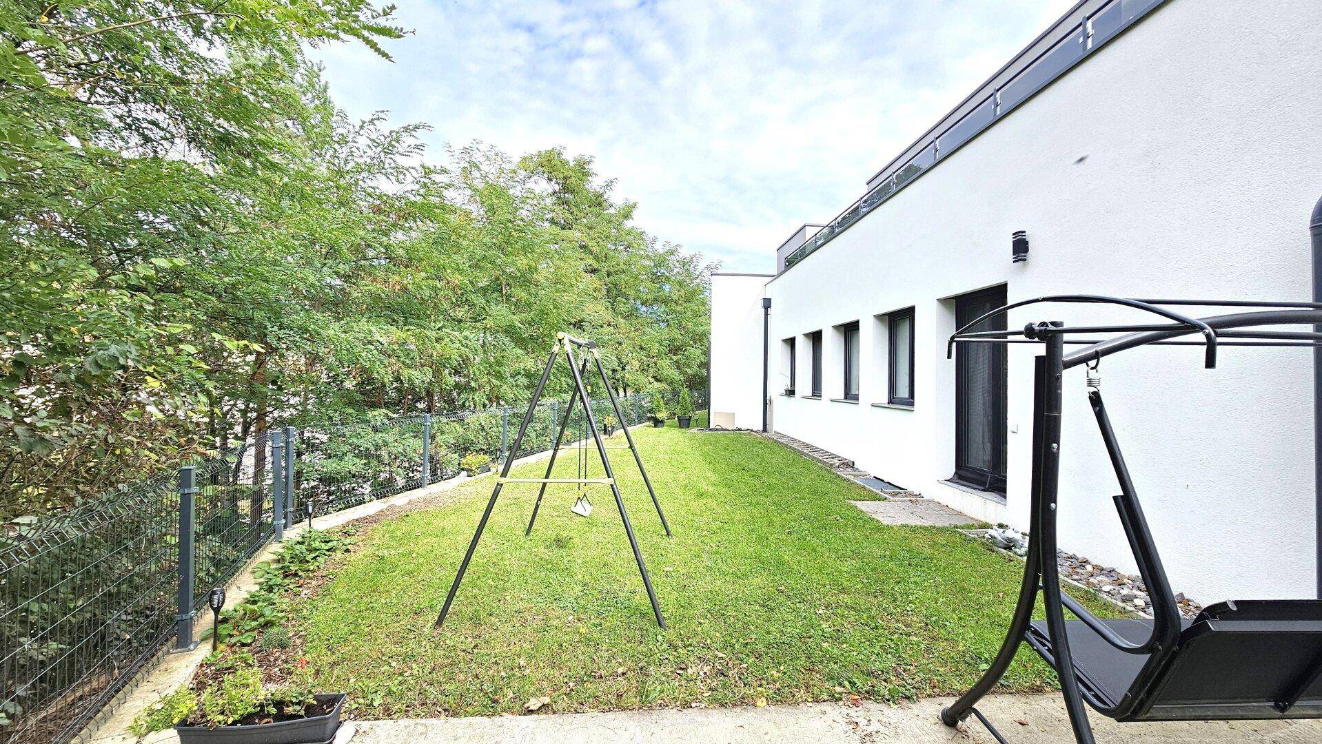 Grüner Gartenbereich mit Rasenfläche, Schaukel und Bäumen im Hintergrund.
