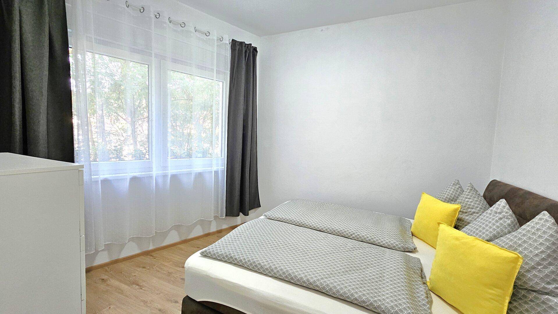 Helles Schlafzimmer mit Doppelbett, Holzboden und dunklen Vorhängen.