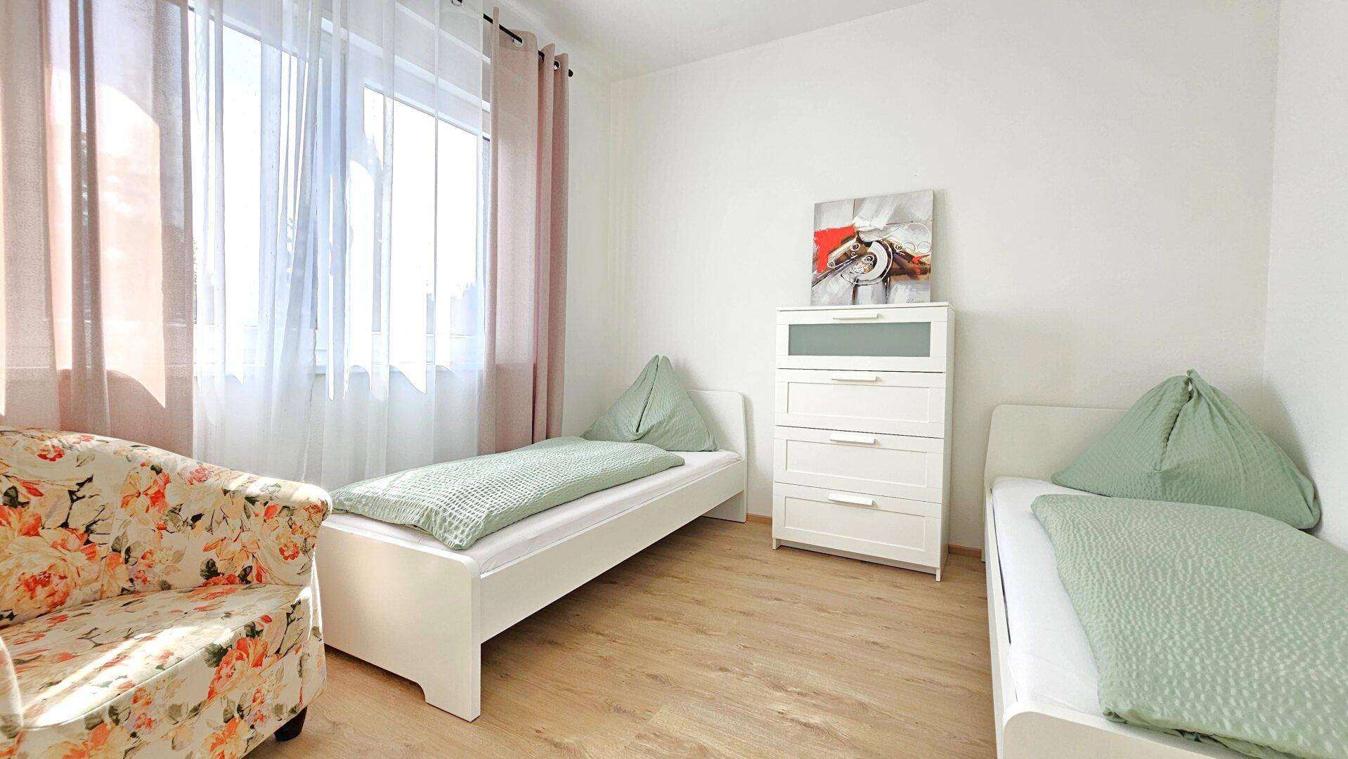 Helles Schlafzimmer mit zwei Einzelbetten, Holzboden und floralem Sessel.