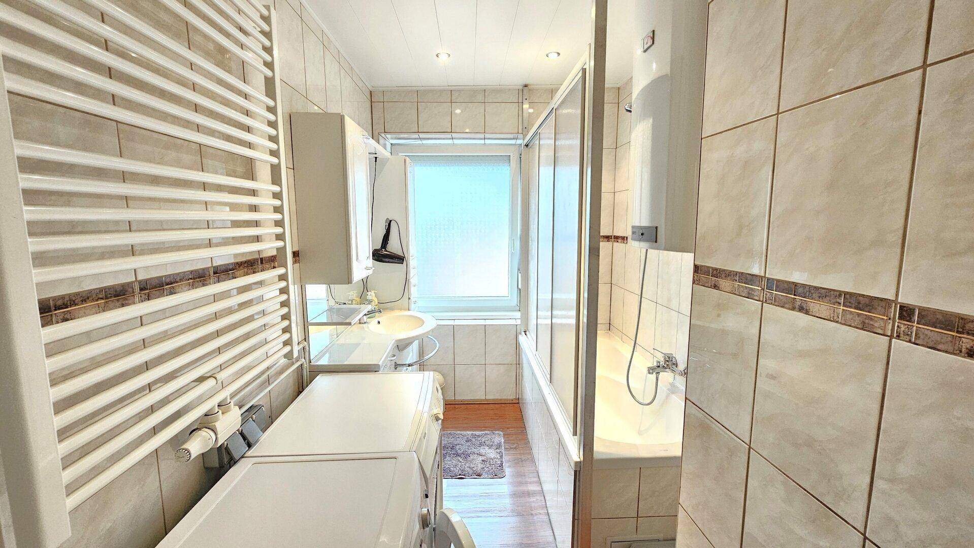 Gefliestes Badezimmer mit Badewanne, Duschwand, Waschbecken und Waschmaschine.