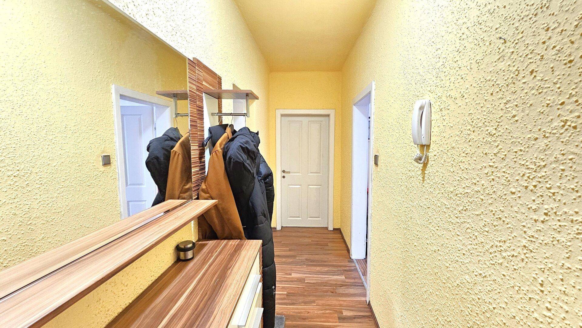 Flur mit gelber Strukturtapete, Holzboden und Garderobe mit Spiegel.