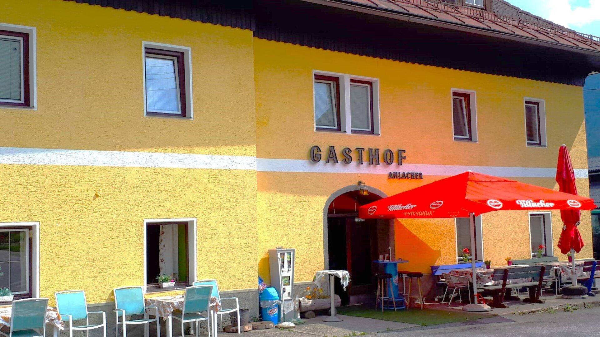 Traditioneller Gasthof mit gelber Fassade, roten Sonnenschirmen und Außenbestuhlung.