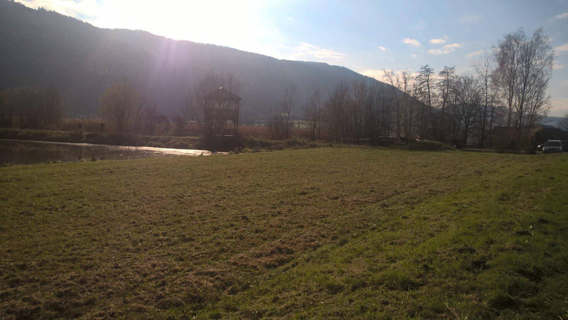Weites Feld mit trockenem Gras, einem Gewässer und einem Aussichtsturm unter strahlendem Himmel.