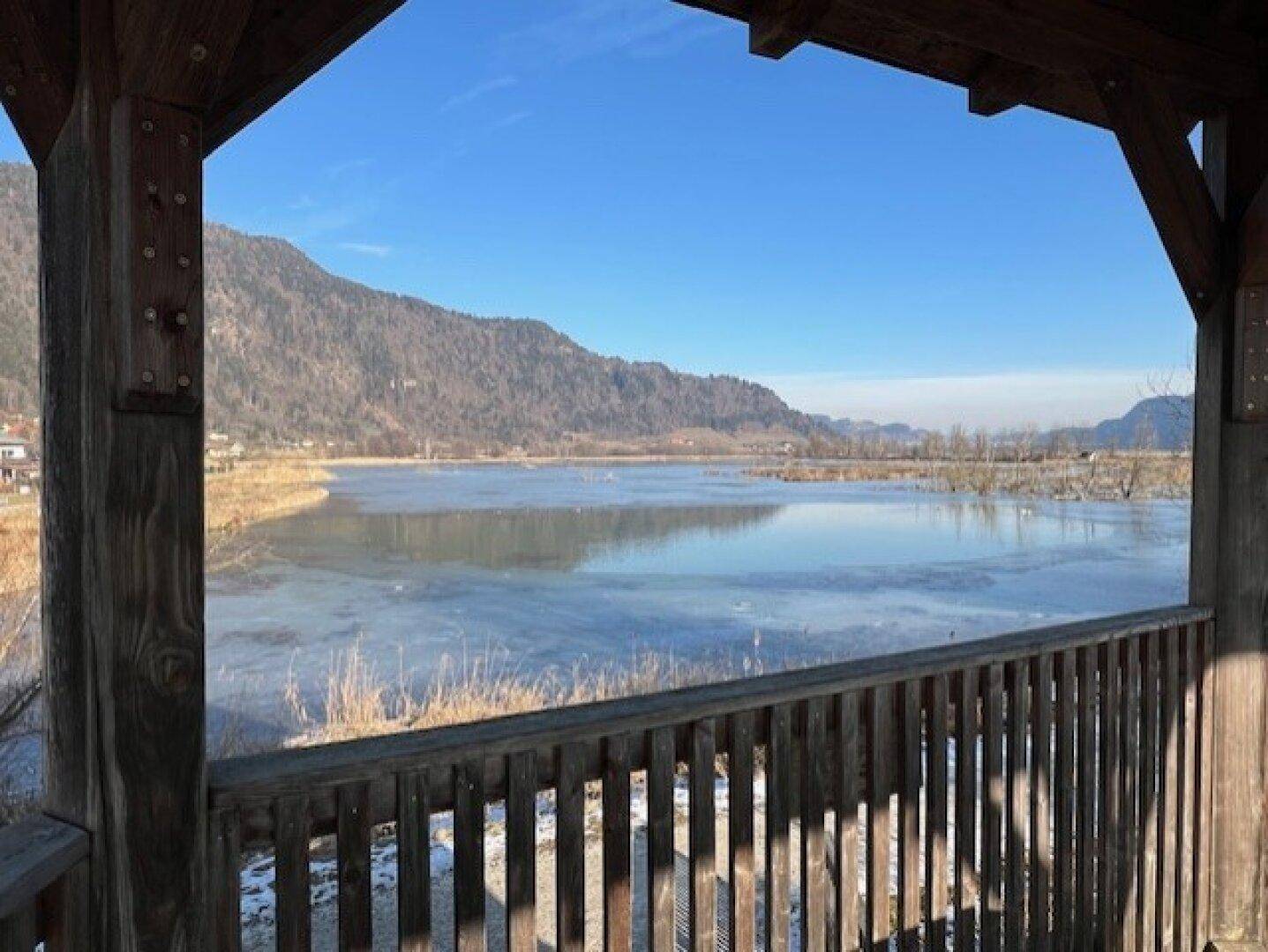 Blick aus einer Holzkonstruktion auf einen teilweise zugefrorenen See und die umliegenden Berge.