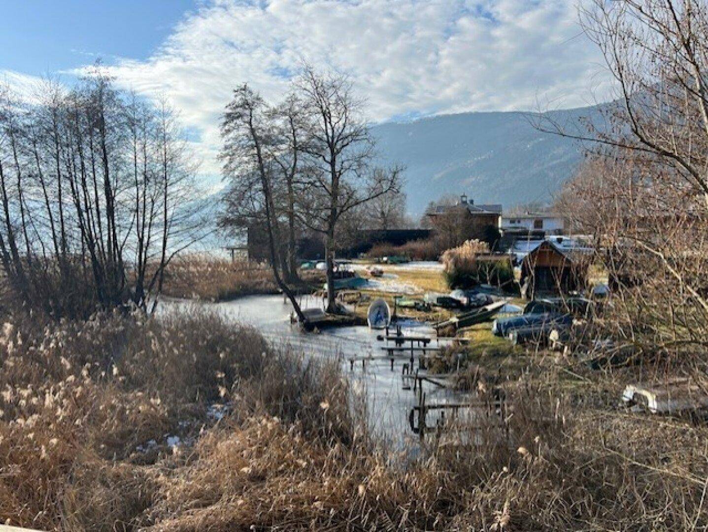 Zugefrorener Bachlauf mit Schilf und Bäumen, der zu einem See führt, umgeben von Bergen.