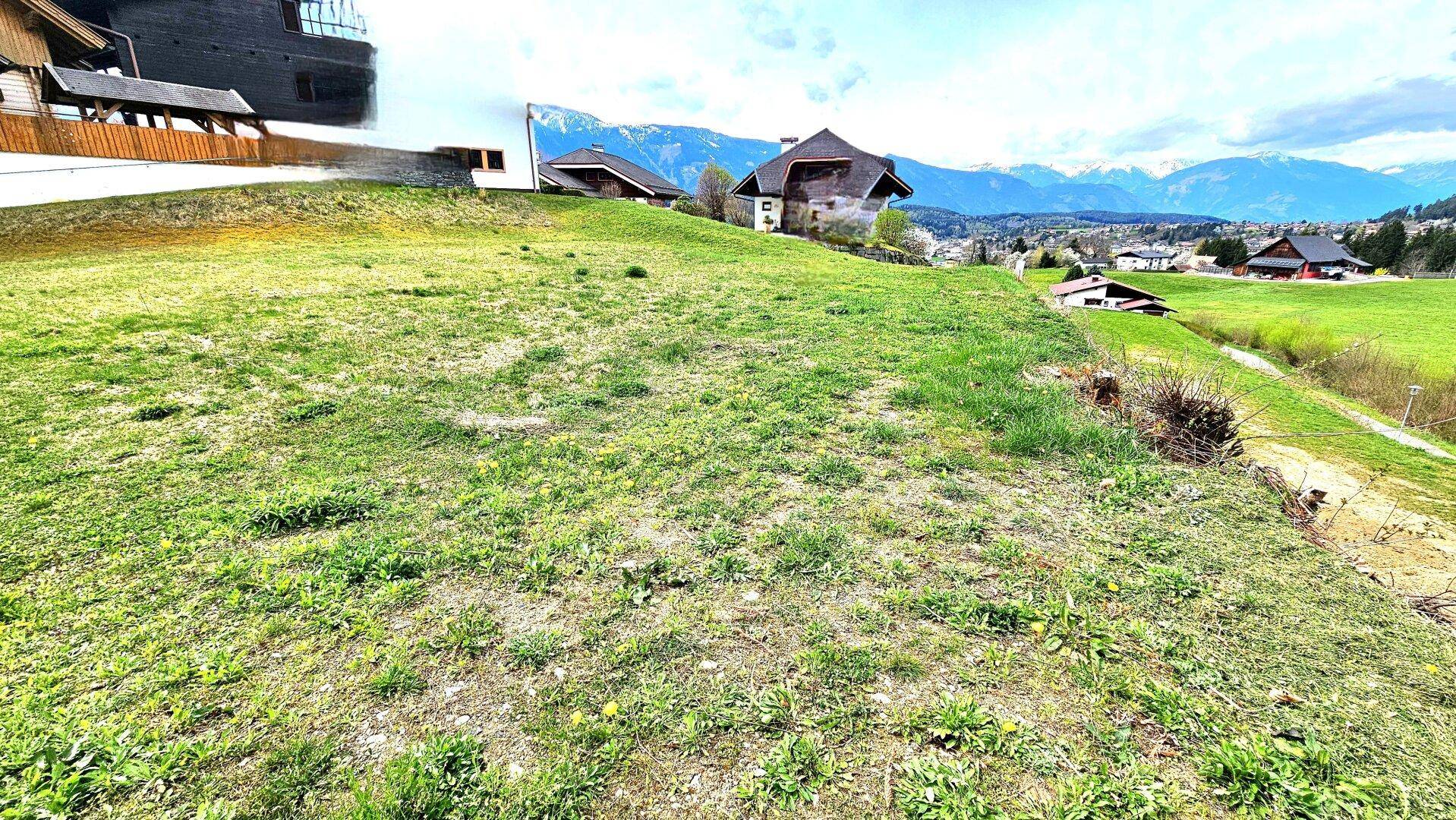 Hanglage mit grüner Wiese und vereinzelten Häusern, umgeben von einer malerischen Berglandschaft.