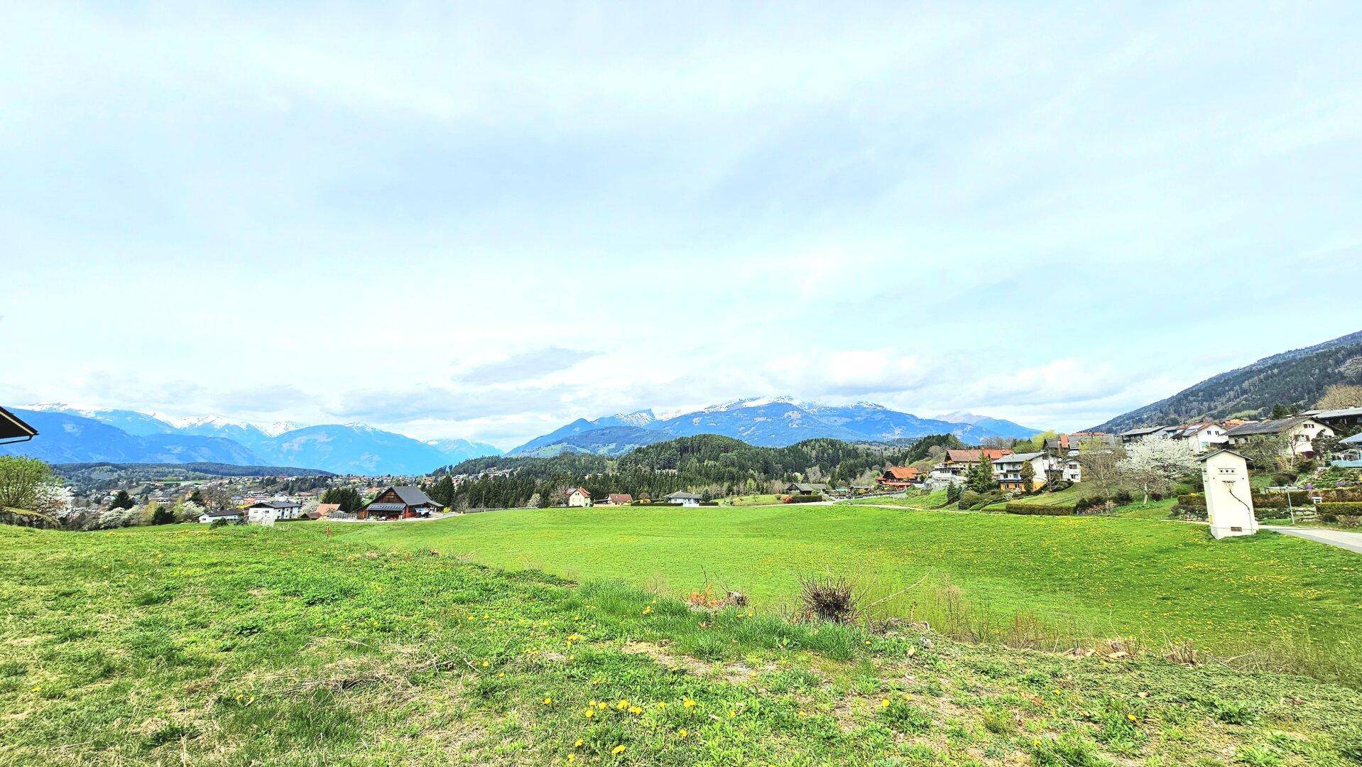 Panoramablick über eine grüne Wiese mit Häusern und schneebedeckten Bergen im weiten Hintergrund.