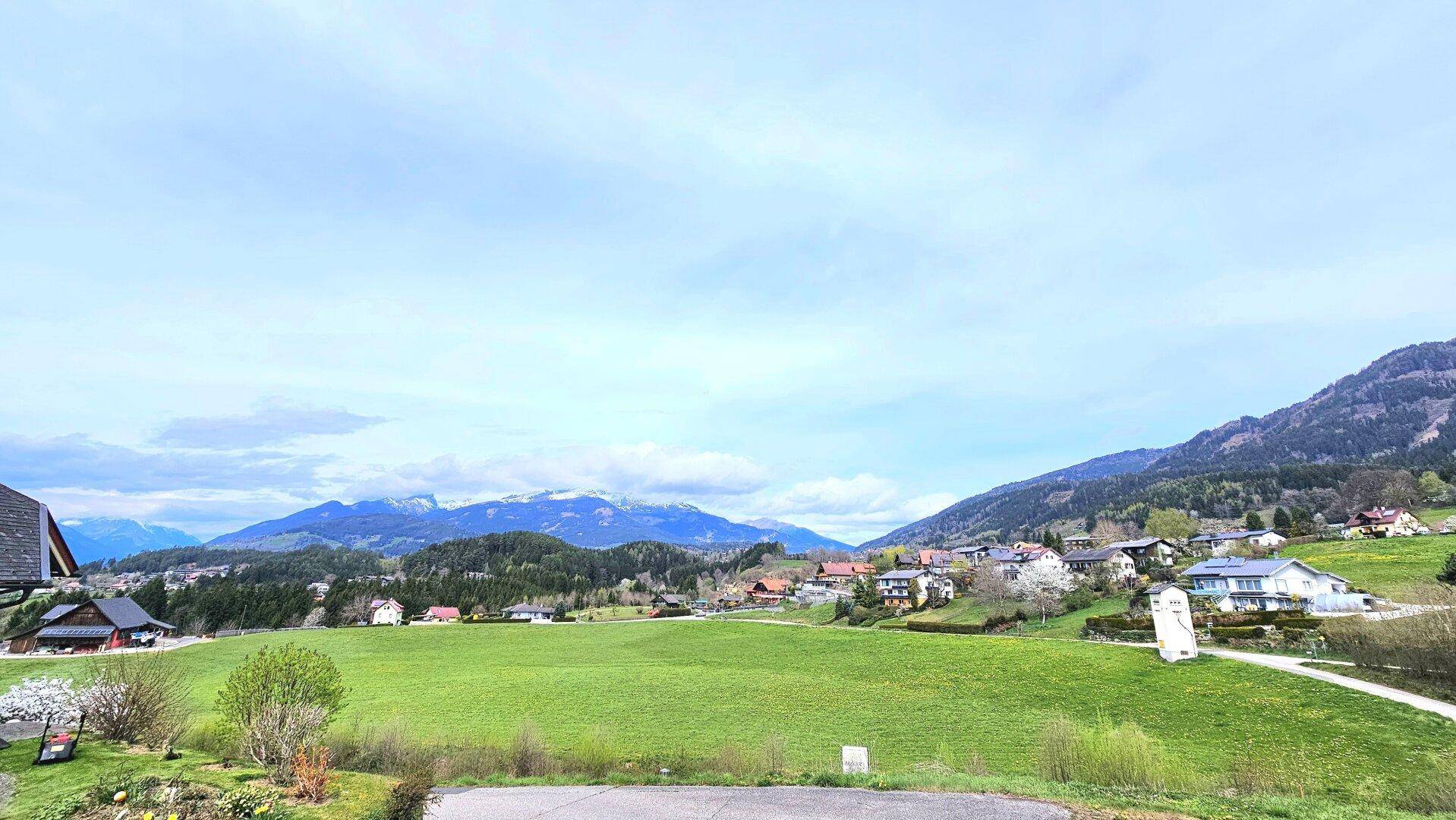 Idyllische Landschaft mit weiten Wiesen, Häusern und einer beeindruckenden Bergkulisse unter blauem Himmel.