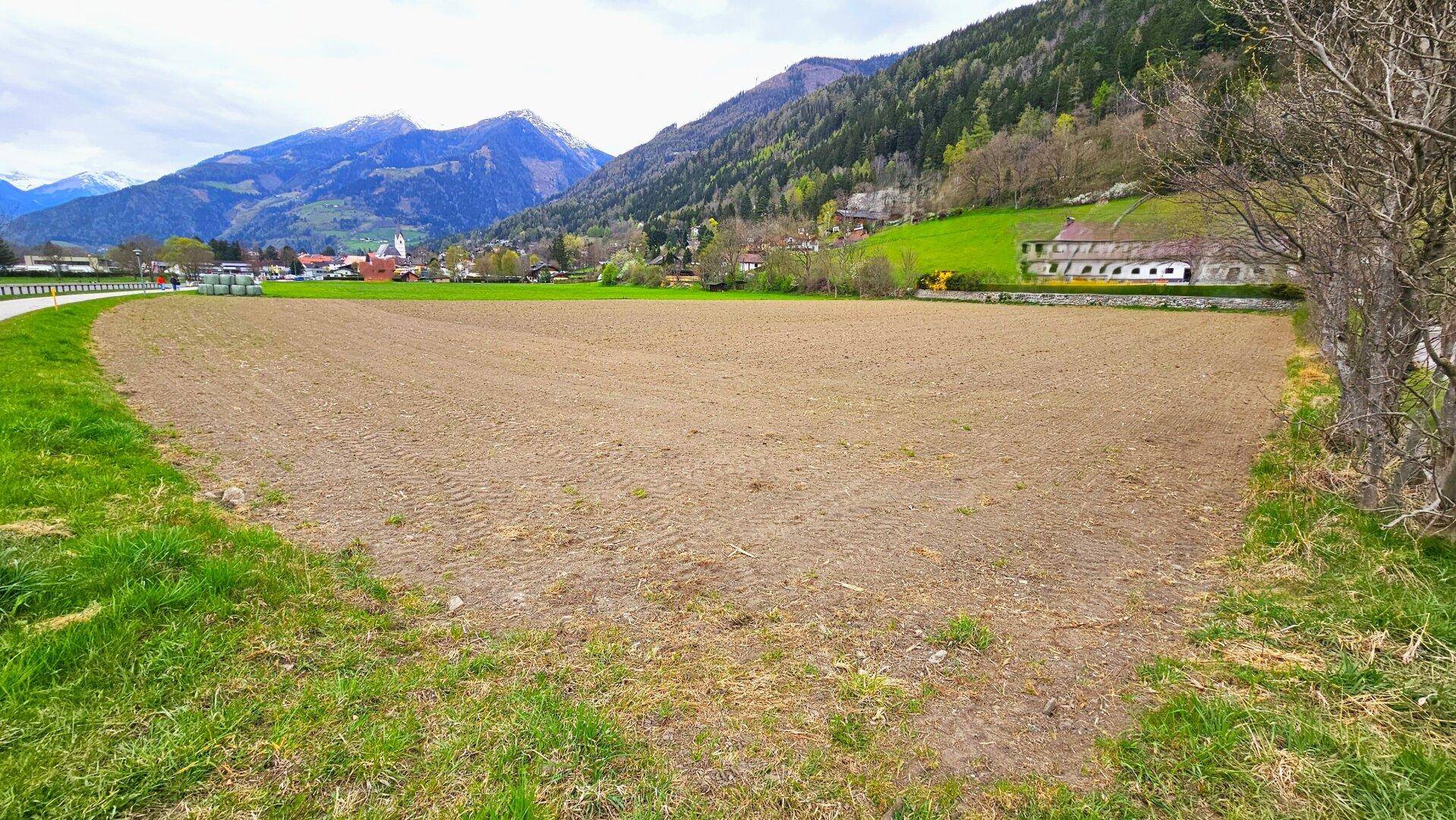 Ackerfläche mit grünen Rändern und einem Bauernhof am Hang, umgeben von Bergen.