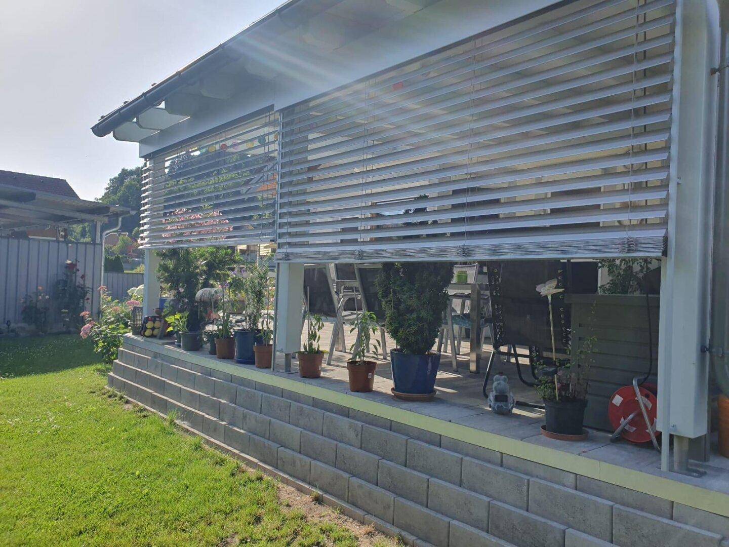 Seitlicher Blick auf die überdachte Terrasse mit Jalousien und Pflanzenarrangements.
