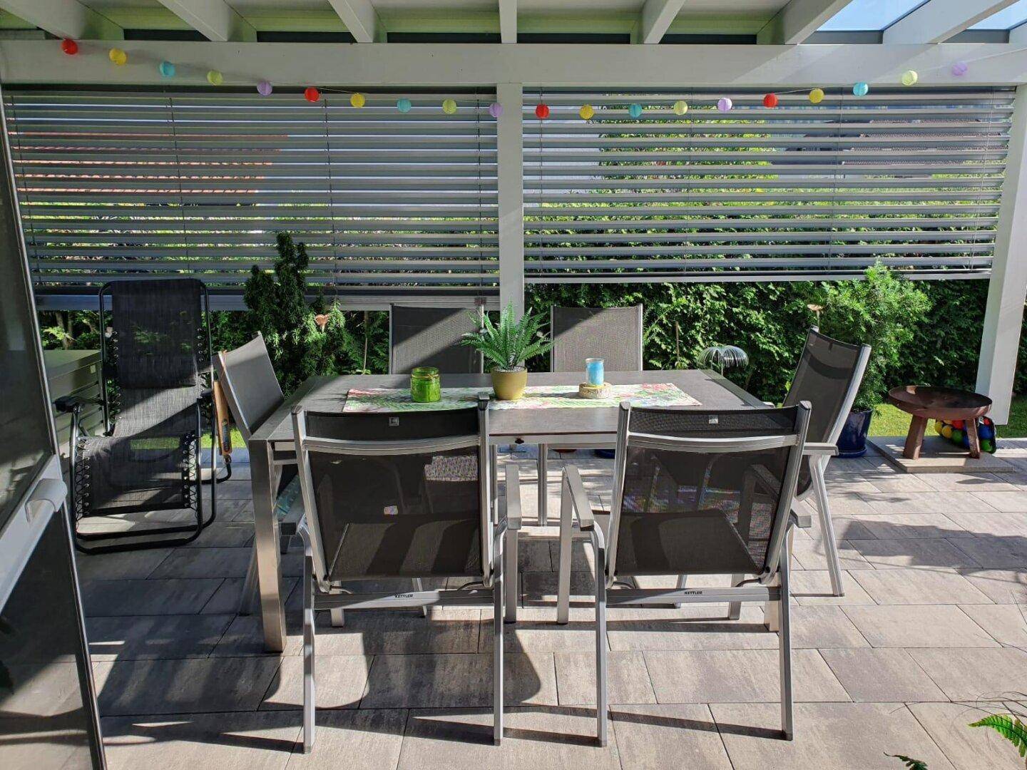 Überdachte Terrasse mit Esstisch, Stühlen und farbenfroher Lichterkette.