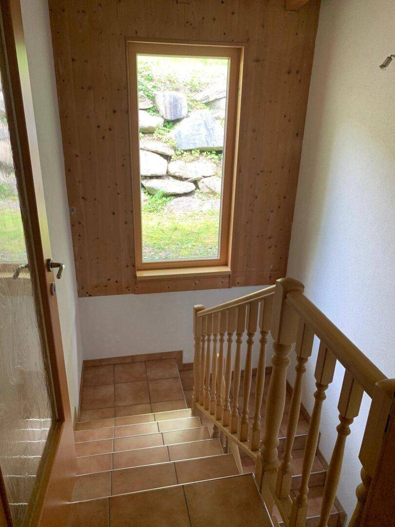 Treppenhaus mit Holzgeländer und Fliesenstufen, bietet Blick auf eine Felswand durch das Fenster.