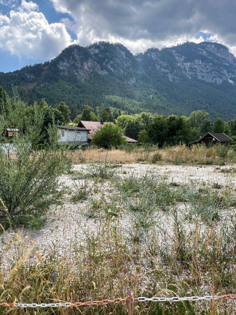 Weitläufiges Gelände mit niedriger Vegetation und Häusern im Hintergrund, umgeben von Bergen.