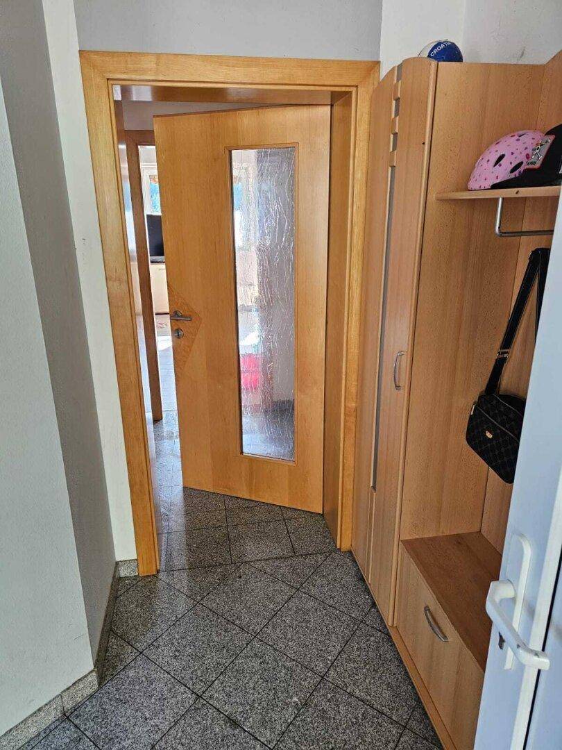 Eingangsbereich mit grauen Fliesen, Holzgarderobe und einer Holztür mit Glaseinsatz.