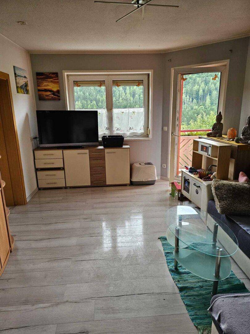 Helles Wohnzimmer mit Laminatboden, TV-Möbeln und Glastisch, mit Zugang zum Außenbereich und Grünblick.