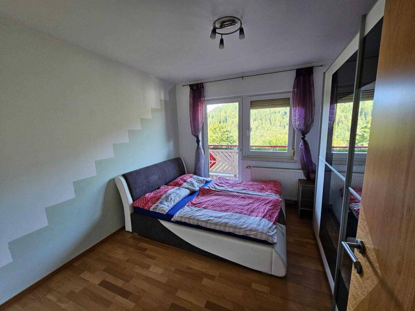 Schlafzimmer mit Bett, Holzboden, Spiegelschrank und Fenster mit Blick ins Grüne.