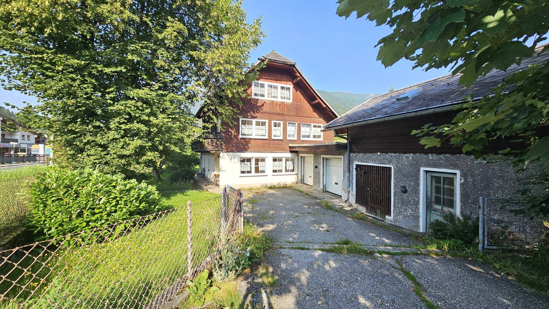 Traditionelles Holzhaus mit Garagen und Zufahrt, umgeben von Bäumen und ländlicher Architektur.