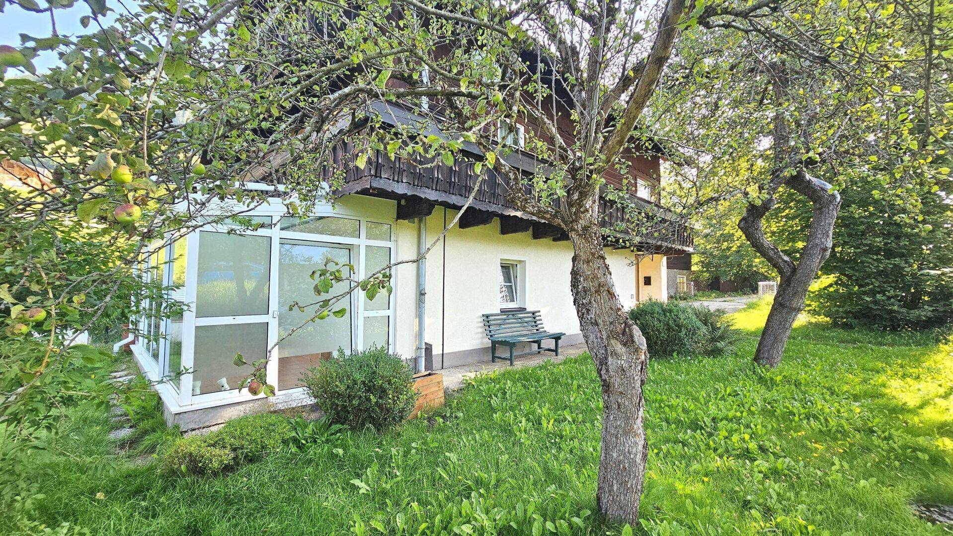Seitlicher Blick auf das Haus mit Wintergarten und Obstbäumen in einem sonnigen Gartenbereich.