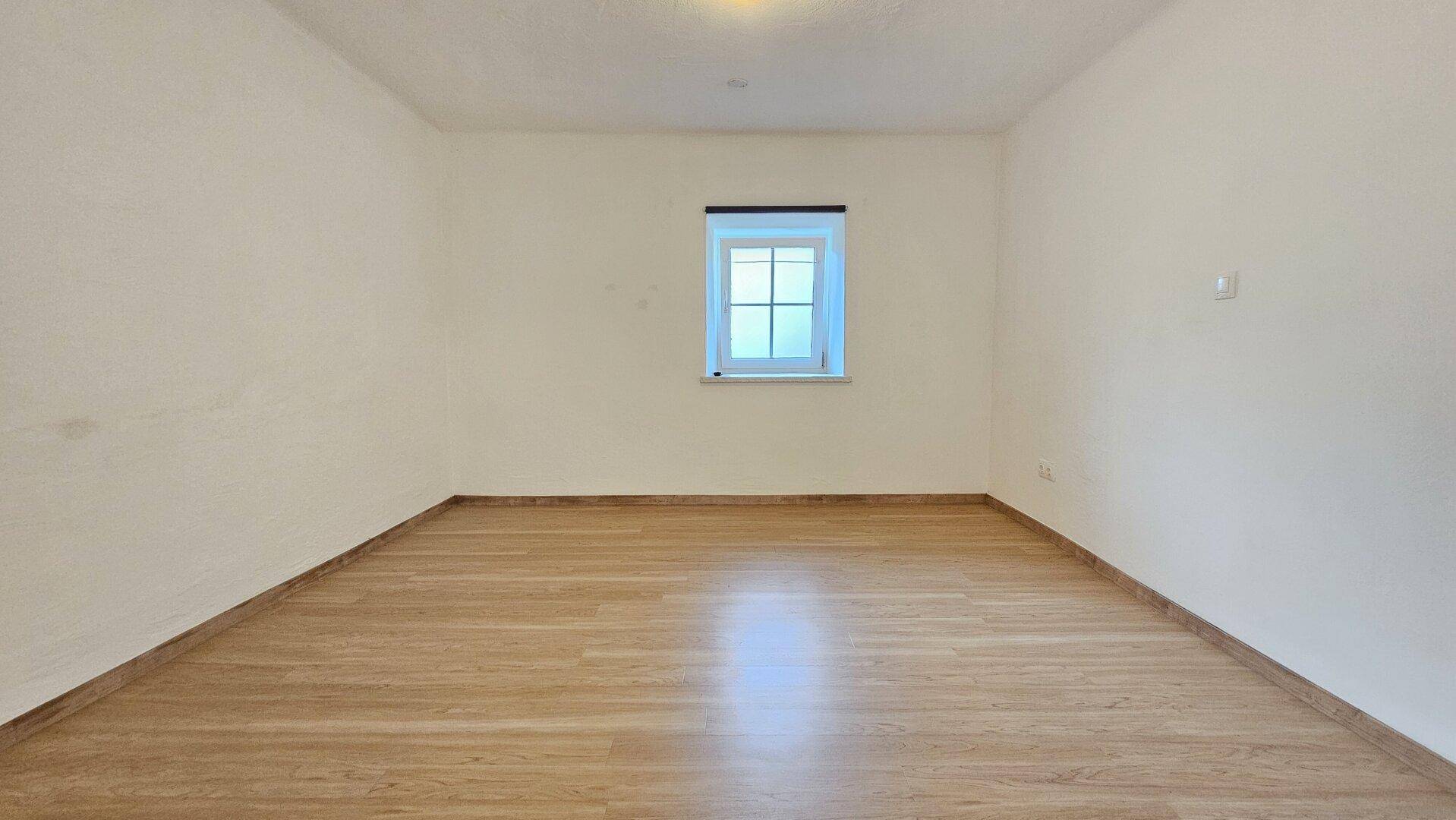 Leeres Zimmer mit hellem Laminatboden und einem Fenster, bereit zur individuellen Gestaltung.